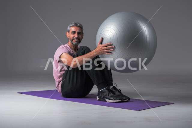 النظر الى الكاميرا بايماءات السعاده ،  نشاط بدني للمحافظة على الصحة و اللياقة الجسدية ، أسلوب حياة صحي ، تقوية عضلات الجسم ، بورتريه لرجل عربي خليجي سعودي يقوم بممارسة التمارين الرياضية مع كرة بيلاتيس ، خلفيه رماديه