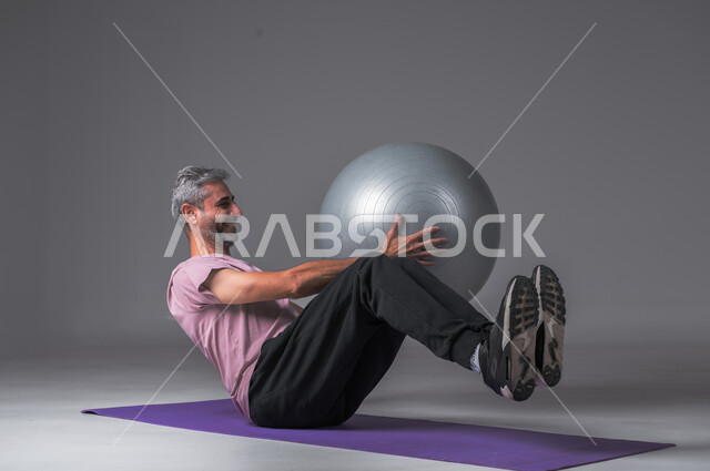 Healthy lifestyle, strengthening body muscles, physical activity to maintain health and fitness, portrait of a Saudi Arabian Gulf man doing exercises with a Pilates ball, gray background