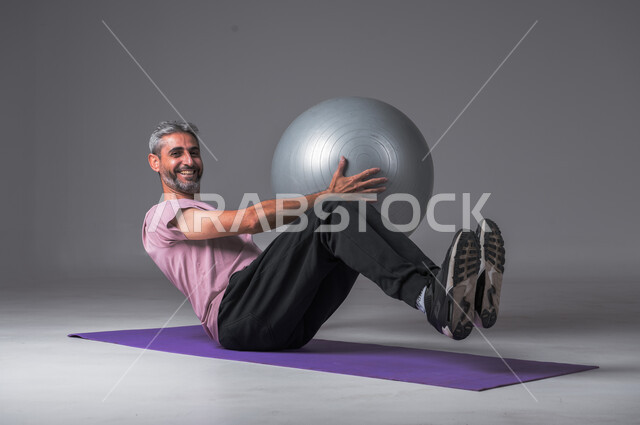 تقوية عضلات الجسم ، نشاط بدني للمحافظة على الصحة و اللياقة الجسدية ، بورتريه لرجل عربي خليجي سعودي يقوم بممارسة التمارين الرياضية مع كرة بيلاتيس ينظر الى الكاميرا بابتسامه ، أسلوب حياة صحي ، خلفيه رماديه