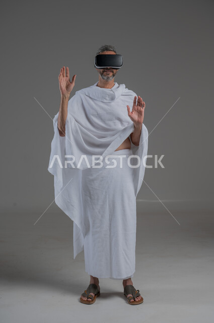 Integration and simulation of the imaginary world, technical and technological development, fun and entertainment by watching through virtual reality glasses, portrait of a Saudi Arabian Gulf man wearing Ihram clothes looking through 3D glasses (VR), full body image, gray background