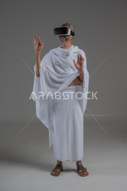 المتعة و التسلية بالمشاهدة عبر نظارة الواقع الافتراضي، التطور التقني والتكنولوجي, بورتريه لرجل عربي خليجي سعودي يرتدي ملابس الاحرام ينظر من خلال نظارات ثلاثية الأبعاد (الفي ار), الاندماج ومحاكاة العالم الخيالي، صورة لكامل الجسم، خلفية رمادية