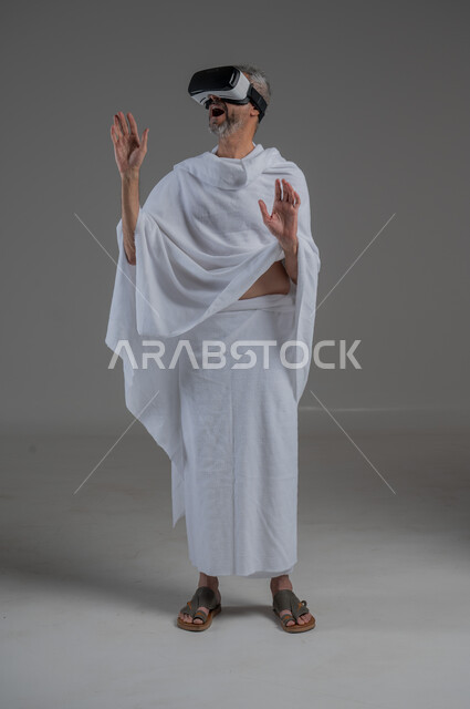 Technical and technological development, fun and entertainment by watching through virtual reality glasses, portrait of a Saudi Arabian Gulf man wearing Ihram clothes looking through 3D glasses (VR), integration and simulation of the imaginary world, full body image, gray background