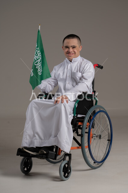 Pride and pride in national identity, Saudi National Day, September 23, portrait of a young Saudi Gulf Arab with special needs wearing casual clothes, holding the flag of the Kingdom of Saudi Arabia in his hand and looking at the camera, Flag Day, March 11, gray background
