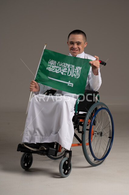 الفخر و الاعتزاز بالهوية الوطنية، اليوم الوطني السعودي 23 سبتمبر، بورتريه لشاب عربي خليجي سعودي من ذوي الهمم يرتدي الزي الكاجوال يحمل بيده علم المملكة العربيه السعودية وينظر إلى الكاميرا، يوم العلم 11 مارس، خلفية رمادية