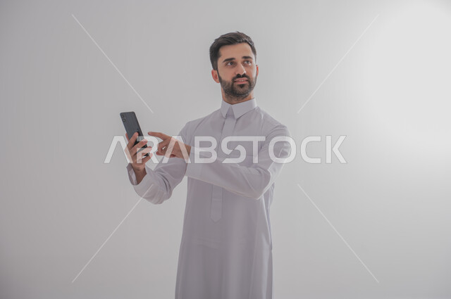 استخدام التكنولوجيا المتطورة للتسويق الرقمي الالكتروني، الإشارة إلى شاشة سوداء فارغة عبر الهاتف المحمول ،عرض التطبيقات والخدمات الحديثة، بورتريه لشاب عربي خليجي سعودي مبتسم يرتدي الثوب التقليدي و يحمل بيده جوال، صورة الجسم بالطول الكامل، خلفية رمادية