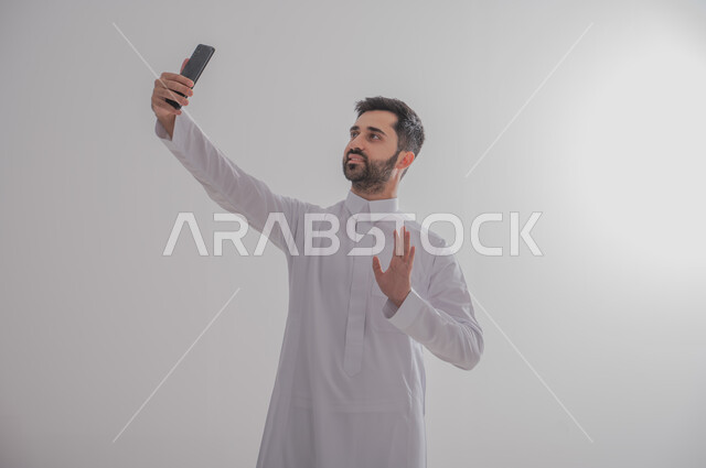 Using advanced technology for electronic digital marketing, pointing to a blank black screen on a mobile phone, displaying modern applications and services, a portrait of a smiling Gulf Arab Saudi man wearing a traditional thobe and holding a mobile phone in his hand while photographing, full-length body photo, gray background