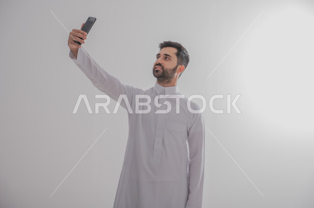 Using advanced technology for electronic digital marketing, pointing to a blank black screen on a mobile phone, displaying modern applications and services, a portrait of a smiling Gulf Arab Saudi man wearing a traditional thobe and holding a mobile phone in his hand while photographing, full-length body photo, gray background