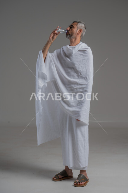 المداومة على شرب المياه، المحافظة على سلامة الجهاز المناعي للجسم، الشعور بالعطش والجفاف، بورتريه من الجانب لرجل عربي خليجي سعودي يرتدي ملابس الاحرام يحمل بيده قارورة من الماء، صورة لكامل الجسم، خلفية رمادية