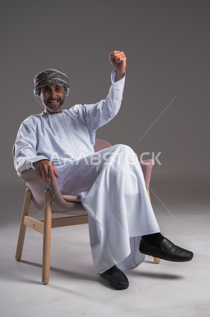 النظر إلى الكاميرا بإيماءات السعادة، الشغف بمتابعة فريق كرة القدم، بورتريه لرجل عربي خليجي عماني يرتدي الدشداشه والعمامة يجلس على كرسي مريح ويتابع التفاز، تعابير وجه ويدين تدل على الحماس والتشجيع، الانتصار وتحقيق الأهداف وفوز الفريق المفضل، خلفية رمادية