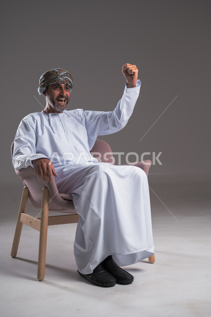 الانتصار وتحقيق الأهداف وفوز الفريق المفضل بالبطولة، الشغف بمتابعة فريق كرة القدم، بورتريه لرجل عربي خليجي عماني يرتدي الدشداشه والعمامة يجلس على كرسي مريح ويتابع التفاز، تعابير وجه ويدين تدل على الحماس والتشجيع، خلفية رمادية