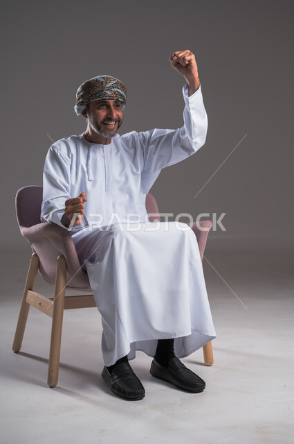 تعابير وجه ويدين تدل على الحماس والتشجيع، الانتصار وتحقيق الأهداف وفوز الفريق المفضل بالبطولة، الشغف بمتابعة فريق كرة القدم، بورتريه لرجل عربي خليجي عماني يرتدي الدشداشه والعمامة يجلس على كرسي مريح ويتابع التفاز، خلفية رمادية