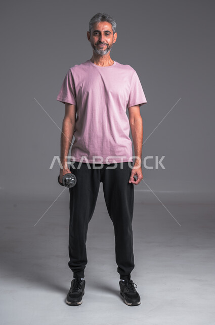 رفع الأوزان الحديدية الثقيلة ، نشاط بدني للمحافظة على الصحة و اللياقة الجسدية ، بورتريه لرجل عربي خليجي سعودي ينظر الى الكاميرا بابتسامه يقوم بممارسة التمارين الرياضية مع الدمبل ، تقوية عضلات الجسم و بنائها ، خلفيه الجسم بالطول الكامل  ، خلفيه رماديه