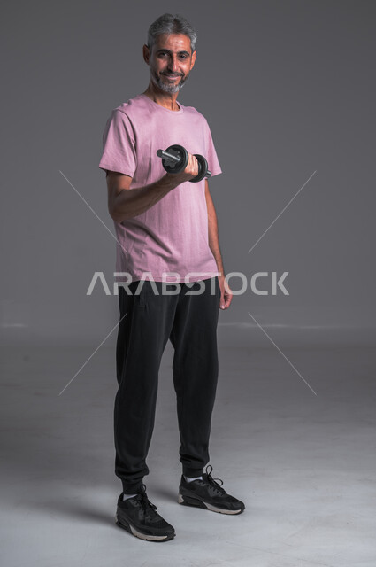  رفع الأوزان الحديدية الثقيلة ، نشاط بدني للمحافظة على الصحة و اللياقة الجسدية ، بورتريه لرجل عربي خليجي سعودي ينظر الى الكاميرا بابتسامه يقوم بممارسة التمارين الرياضية مع الدمبل ، تقوية عضلات الجسم و بنائها ، صوره الجسم بالطول الكامل ، خلفيه رماديه