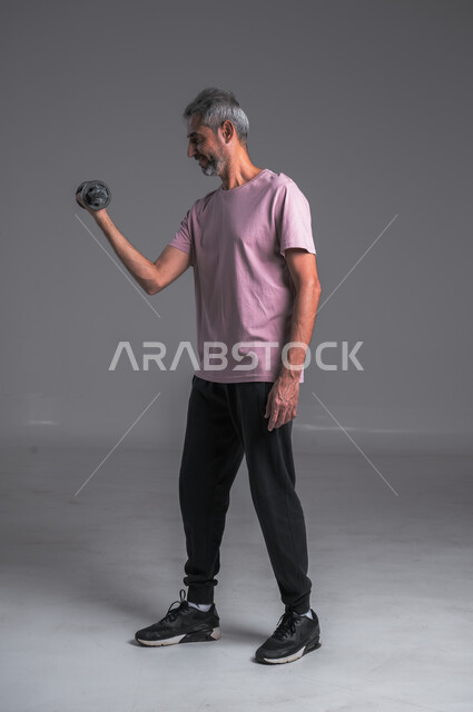 تقوية عضلات الجسم و بنائها ، نشاط بدني للمحافظة على الصحة و اللياقة الجسدية ، بورتريه لرجل عربي خليجي سعودي يقوم بممارسة التمارين الرياضية مع الدمبل ،رفع الأوزان الحديدية الثقيلة ، صوره الجسم بالطول الكامل ، خلفيه رماديه