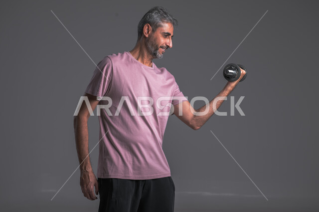  نشاط بدني للمحافظة على الصحة و اللياقة الجسدية ،تقوية عضلات الجسم و بنائها ، رفع الأوزان الحديدية الثقيلة ، بورتريه لرجل عربي خليجي سعودي يقوم بممارسة التمارين الرياضية مع الدمبل ، خلفيه رماديه