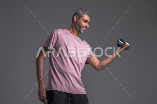 Strengthening and building body muscles, lifting heavy iron weights, physical activity to maintain health and physical fitness, portrait of a Saudi Arabian Gulf man doing exercises with dumbbells, gray background
