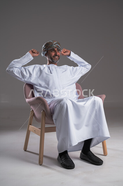 ايماءات وجه ويدين تدل على الراحة، النظر الى شيء ما بتعابير التفكيروشرود الذهن، الشعور بالتعب والحاجة إلى النوم، مفهوم الاسترخاء والاستمتاع بالأجواء الهادئة، بورتريه لرجل عربي خليجي عماني يرتدي الدشداشه والعمامة يجلس على كرسي مريح، خلفية رمادية 