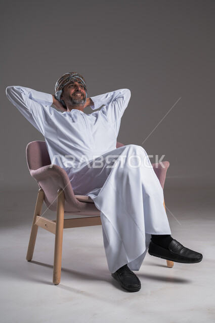 الشعور بالتعب والحاجة إلى النوم، مفهوم الاسترخاء والاستمتاع بالأجواء الهادئة، بورتريه لرجل عربي خليجي عماني يرتدي الدشداشه والعمامة يجلس على كرسي مريح ويضع يديه تحت رأسه، تعابير وإيماءات الراحة والشرود، النظر الى شيء ما، خلفية رمادية 