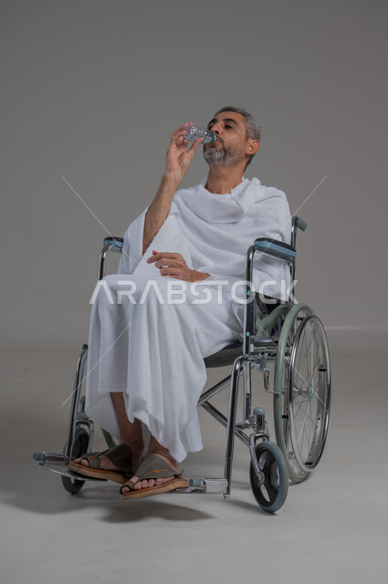 الشعور بالعطش والجفاف، المحافظة على سلامة الجهاز المناعي للجسم، المداومة على شرب المياه، بورتريه من الجانب لرجل عربي خليجي سعودي مقعد يرتدي ملابس الاحرام يجلس على كرسي متحرك يحمل بيده قارورة من الماء، صورة لكامل الجسم، خلفية رمادية