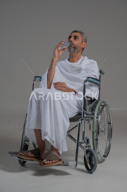 المحافظة على سلامة الجهاز المناعي للجسم، المداومة على شرب المياه، الشعور بالعطش والجفاف، بورتريه من الجانب لرجل عربي خليجي سعودي مقعد يرتدي ملابس الاحرام يجلس على كرسي متحرك يحمل بيده قارورة من الماء، صورة لكامل الجسم، خلفية رمادية