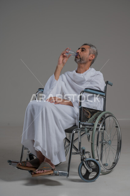 المداومة على شرب المياه، المحافظة على سلامة الجهاز المناعي للجسم، الشعور بالعطش والجفاف، بورتريه من الجانب لرجل عربي خليجي سعودي مقعد يرتدي ملابس الاحرام يجلس على كرسي متحرك يحمل بيده قارورة من الماء، صورة لكامل الجسم، خلفية رمادية