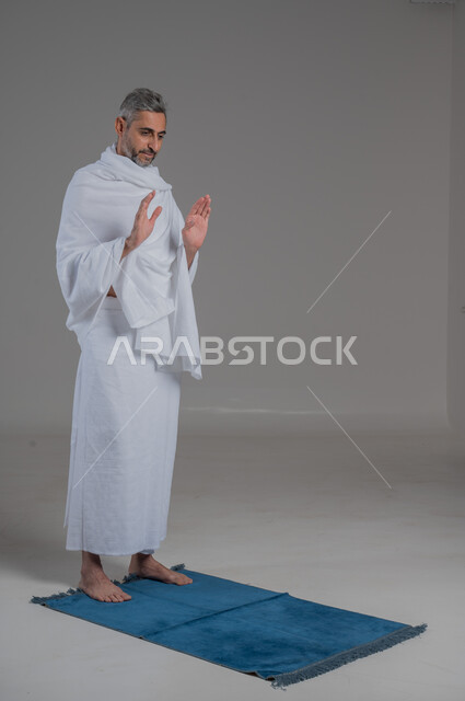قضاء مناسك الحج والعمرة، العبادة والتقرب إلى الله، أداء فرائض الصلاة بأوقاتها، مفهوم أركان الإسلام، بورتريه لرجل عربي خليجي سعودي يرتدي لباس الاحرام يقف على السجادة، موسم الحج 2024، صورة لكامل الجسم، خلفية رمادية