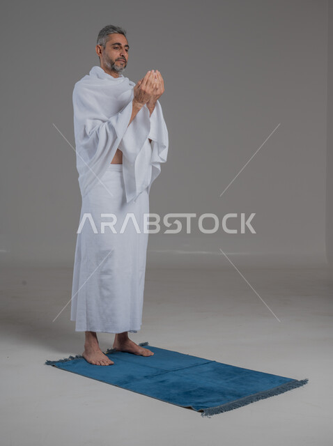 Performing the obligatory prayers on time, supplication and supplication to the Lord of the servants, the concept of the pillars of Islam, a portrait of a Saudi Arabian Gulf man wearing the Ihram clothing standing on the prayer rug, worship and drawing closer to God, Hajj season 2024, a full body image, gray background