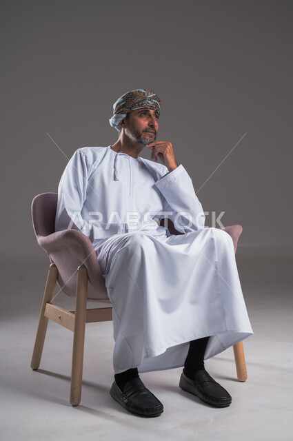 Meditation and planning for the future, finding a solution to a problem and making decisions, looking at something with expressions of thinking and absent-mindedness, portrait of an Arab Gulf Omani man wearing a dishdasha and turban sitting on a comfortable chair and placing his hands under his chin, gray background