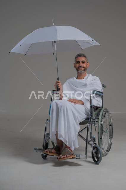 زيارة بيت الله الحرام في مكة المكرمة، ايماءات البهجة والسرور، اداء مناسك الحج والعمرة، بورتريه لرجل عربي خليجي سعودي مقعد يرتدي ملابس الاحرام يجلس على كرسي متحرك يحمل بيده مظلة للحماية من أشعة الشمس، صورة لكامل الجسم، خلفية رمادية