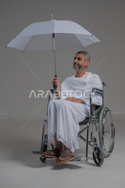 ايماءات البهجة والسرور، زيارة بيت الله الحرام في مكة المكرمة، اداء مناسك الحج والعمرة، بورتريه لرجل عربي خليجي سعودي مقعد يرتدي ملابس الاحرام يجلس على كرسي متحرك يحمل بيده مظلة للحماية من أشعة الشمس، صورة لكامل الجسم، خلفية رمادية