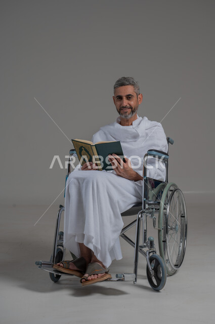 قراءة كتاب الله وحفظه وتجويده، تلاوة المصحف الشريف، مفهوم الاجتهاد في العبادة، اداء مناسك الحج والعمرة، بورتريه لرجل عربي خليجي سعودي مقعد يرتدي ملابس الاحرام يجلس على كرسي متحرك يحمل بيده القرأن، صورة لكامل الجسم، خلفية رمادية