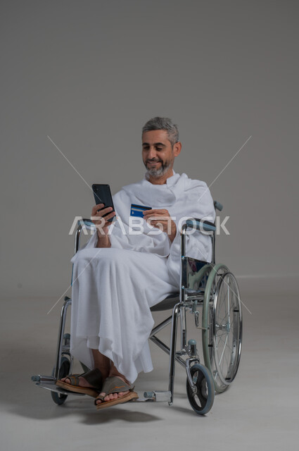 الالكتروني في حملات الحج والعمرة، الشراء والدفع عن طريق بطاقة الصراف الإئتمانية, الحجز بورتريه لرجل عربي خليجي سعودي مقعد يرتدي ملابس الاحرام يجلس على كرسي متحرك يحمل بيده الهاتف الجوال وكرت البنك المصرفي, حجوزات الاونلاين السريعة، صورة لكامل الجسم، خلفية رمادية