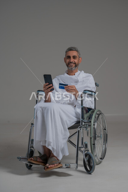 الشراء والدفع عن طريق بطاقة الصراف الإئتمانية, الحجز الالكتروني في حملات الحج والعمرة، بورتريه لرجل عربي خليجي سعودي مقعد يرتدي ملابس الاحرام يجلس على كرسي متحرك يحمل بيده الهاتف الجوال وكرت البنك المصرفي, حجوزات الاونلاين السريعة، صورة لكامل الجسم، خلفية رمادية