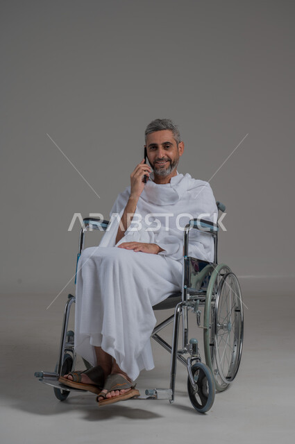 التواصل مع الاهل والاصدقاء، اجراء مكالمات هاتفية، ايماءات السرور و السعادة، استخدام الاجهزة التقنية الحديثة، بورتريه لرجل عربي خليجي سعودي مقعد يرتدي ملابس الاحرام يجلس على كرسي متحرك يحمل بيده الهاتف المحمول, صورة لكامل الجسم، خلفية رمادية