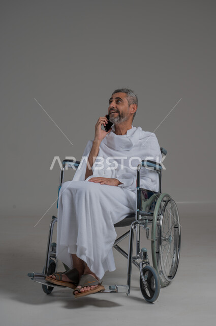 Gestures of pleasure and happiness, making phone calls, using modern technical devices, communicating with family and friends, portrait of a disabled Saudi Arabian Gulf man wearing Ihram clothes sitting on a wheelchair holding a mobile phone in his hand, full body image, gray background