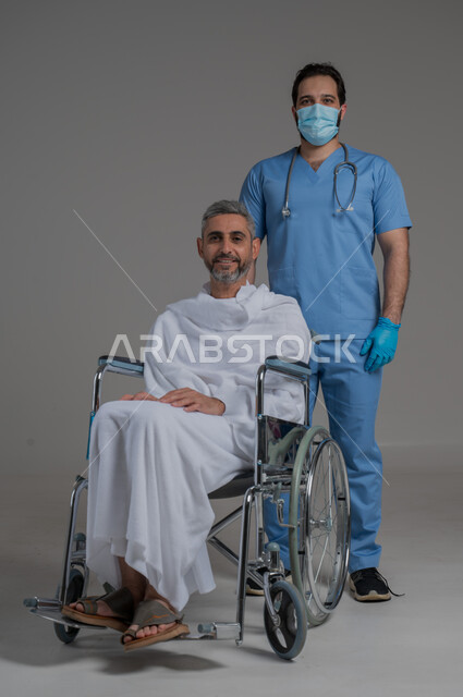 الاهتمام بصحة المعتمرين، أداء مناسك الحج والعمرة، تقديم الخدمات الطبيه لحجاج بيت الله الحرام، بورتريه لممرض اسعافات اولية يرتدي اليونيفورم والكمامة يقوم بالاهتمام برجل عربي خليجي سعودي مقعد يرتدي ملابس الاحرام يجلس على كرسي متحرك, صورة لكامل الجسم، خلفية رمادية