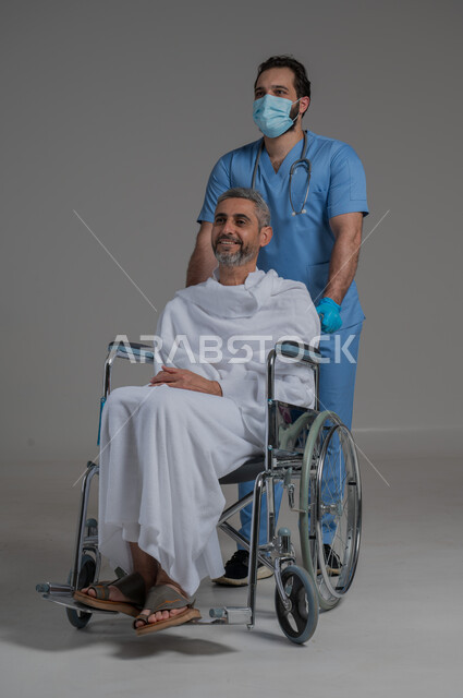 أداء مناسك الحج والعمرة، تقديم الخدمات الطبيه لحجاج بيت الله الحرام، الاهتمام بصحة المعتمرين، بورتريه لممرض اسعافات اولية يرتدي اليونيفورم والكمامة يقوم بالاهتمام برجل عربي خليجي سعودي مقعد يرتدي ملابس الاحرام يجلس على كرسي متحرك, صورة لكامل الجسم، خلفية رمادية
