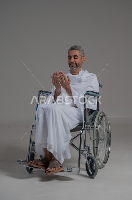 العبادة والتقرب الى الله، اداء مناسك الحج والعمرة، مفهوم أركان الإسلام، التضرع لرب العباد, بورتريه لرجل عربي خليجي سعودي مقعد يرتدي ملابس الاحرام يجلس على كرسي متحرك يرفع يديه بايماءات الدعاء, صورة لكامل الجسم، خلفية رمادية
