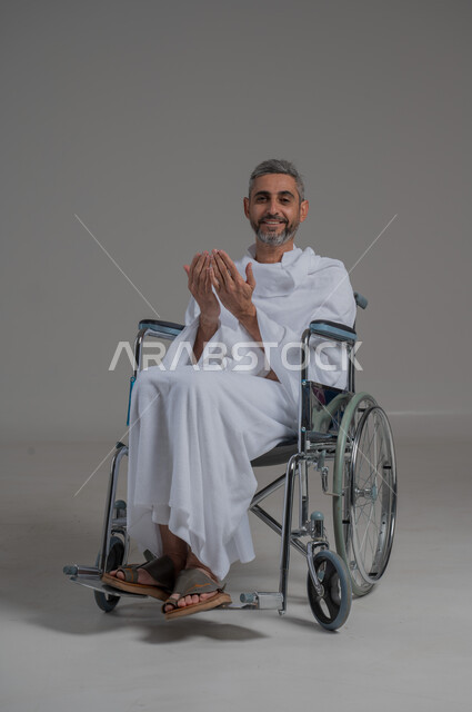 اداء مناسك الحج والعمرة، مفهوم أركان الإسلام، التضرع لرب العباد, العبادة والتقرب الى الله، بورتريه لرجل عربي خليجي سعودي مقعد يرتدي ملابس الاحرام يجلس على كرسي متحرك يرفع يديه بايماءات الدعاء, صورة لكامل الجسم، خلفية رمادية