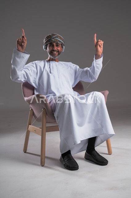 تحديد موقع معين بايماءات السعادة والسرور، إشارات وحركات يدين تدل على هذا المكان (هنا)، بورتريه لرجل عربي خليجي عماني يرتدي الدشداشه والعمامه يجلس على كرسي مريح يرفع اصبعه السبابة إلى الأعلى، النظر والإشارة إلى شيء ما، خلفية رمادية