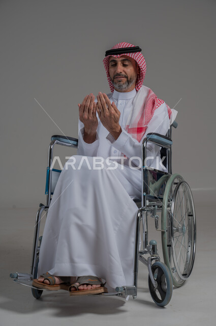 التضرع لرب العباد, العبادة والتقرب الى الله، مفهوم أركان الإسلام، بورتريه لرجل عربي خليجي سعودي مقعد يرتدي الثوب التقليدي والشماغ يجلس على كرسي متحرك يرفع يديه بايماءات الدعاء, صورة لكامل الجسم، خلفية رمادية