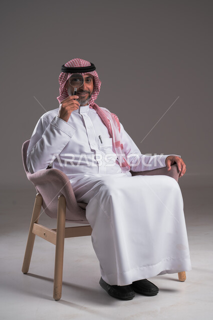 مفهوم توضيح الصورة، ايماءات تدل على السعادة والسرور، بورتريه لرجل عربي خليجي سعودي مبتسم يرتدي الثوب التقليدي والشماغ يجلس على كرسي مريح وينظر من خلال العدسة المكبرة، الفحص والاستكشاف والرؤية عن قرب، التدقيق والتركيز باستخدام الأدوات البصرية، خلفية رمادية