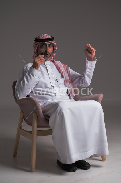  الفحص والاستكشاف والرؤية عن قرب، التدقيق والتركيز باستخدام الأدوات البصرية، بورتريه لرجل عربي خليجي سعودي يرتدي الثوب التقليدي والشماغ يجلس على كرسي مريح وينظر من خلال العدسة المكبرة، مفهوم توضيح الصورة، ايماءات وجه ويدين تدل على السعادة والدهشة، خلفية رمادية
