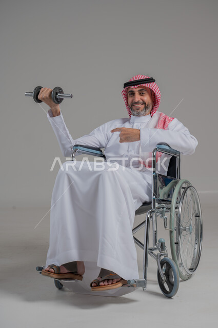 مفهوم الاهتمام بذوي الاحتياجات الخاصة، تنمية ودعم القدرة الحركية على حمل ورفع الأشياء، بورتريه لرجل عربي خليجي سعودي مقعد يرتدي الثوب التقليدي والشماغ يجلس على كرسي متحرك يحمل بيده دمبلز، صورة لكامل الجسم، خلفية رمادية