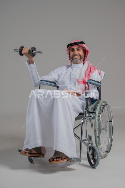 تنمية ودعم القدرة الحركية على حمل ورفع الأشياء، مفهوم الاهتمام بذوي الاحتياجات الخاصة، بورتريه لرجل عربي خليجي سعودي مقعد يرتدي الثوب التقليدي والشماغ يجلس على كرسي متحرك يحمل بيده دمبلز، صورة لكامل الجسم، خلفية رمادية