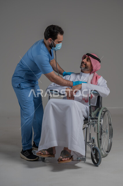 ايماءات السرور والسعادة، مفهوم الرعاية الصحية والاعتناء بالمرضى والمقعدين، نمط حياة ذوي الهمم، بورتريه لممرض يرتدي اليونيفورم والكمامة يقوم بالاعتناء برجل عربي خليجي سعودي مقعد يرتدي الثوب التقليدي والشماغ يجلس على كرسي متحرك، صورة لكامل الجسم، خلفية رمادية