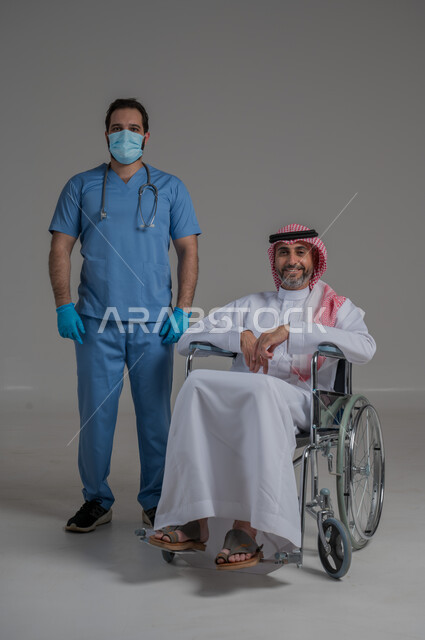 ايماءات السرور والسعادة، مفهوم الرعاية الصحية والاعتناء بالمرضى والمقعدين، نمط حياة ذوي الهمم، بورتريه لممرض يرتدي اليونيفورم والكمامة يقوم بالاعتناء برجل عربي خليجي سعودي مقعد يرتدي الثوب التقليدي والشماغ يجلس على كرسي متحرك، صورة لكامل الجسم، خلفية رمادية