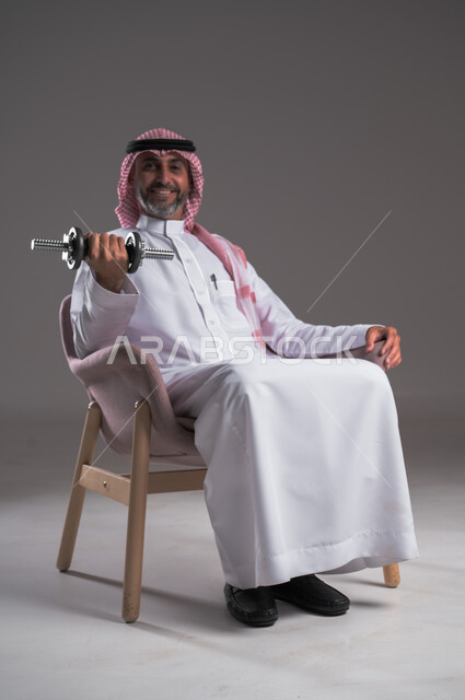 التحفيز والقوة والتحدي لأسلوب حياة صحي، تمارين رفع الأثقال والأوزان الحديدية الثقيلة، بورتريه لرجل عربي خليجي سعودي مبتسم يرتدي الثوب التقليدي والشماغ يجلس على كرسي مريح ويمارس التمارين الرياضية بواسطة الدامبلز، تقوية عضلات الجسم وبناؤها، خلفية رمادية