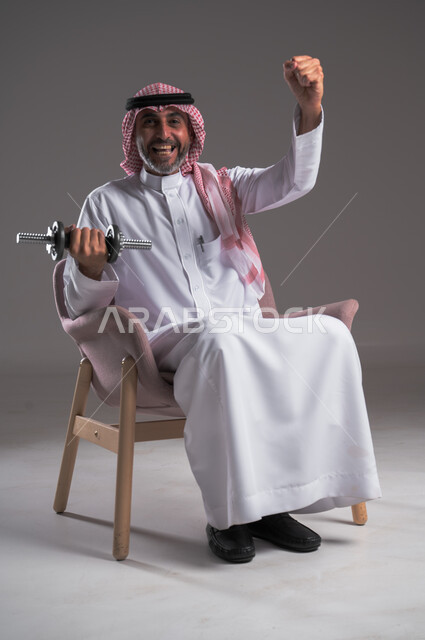 القوة والتحدي والتحفيز لأسلوب حياة صحي، بورتريه لرجل عربي خليجي سعودي مبتسم يرتدي الثوب التقليدي والشماغ يجلس على كرسي مريح ويمارس التمارين الرياضية بواسطة الدامبلز، تقوية عضلات الجسم وبناؤها، تمارين رفع الأثقال والأوزان الحديدية الثقيلة، خلفية رمادية