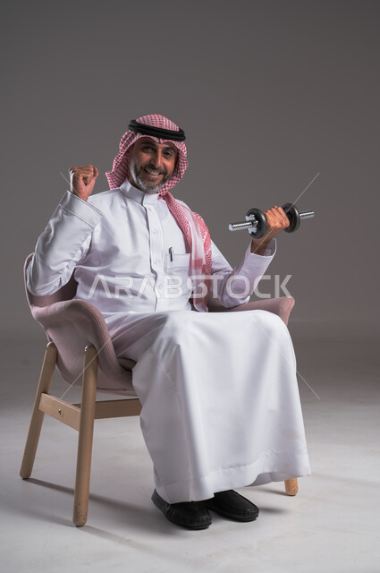 تمارين رفع الأثقال والأوزان الحديدية الثقيلة، القوة والتحدي والتحفيز لأسلوب حياة صحي، بورتريه لرجل عربي خليجي سعودي مبتسم يرتدي الثوب التقليدي والشماغ يجلس على كرسي مريح ويمارس التمارين الرياضية بواسطة الدامبلز، تقوية عضلات الجسم وبناؤها، خلفية رمادية